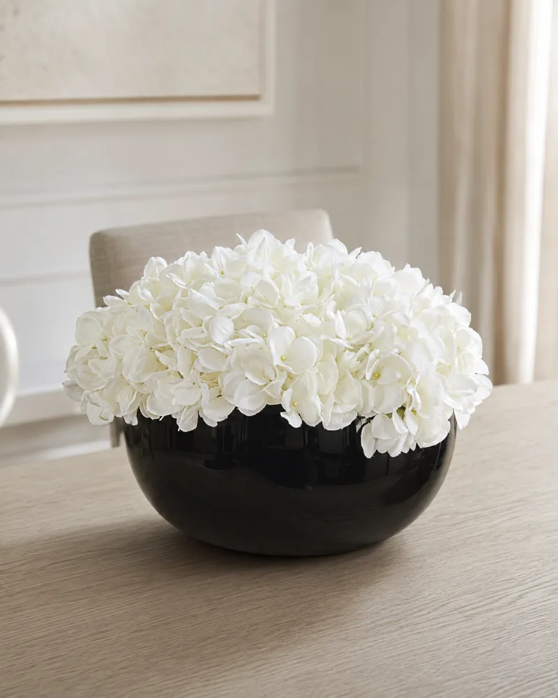 Glossy black bowl holding a dome of white silk hydrangeas on an oak dining table