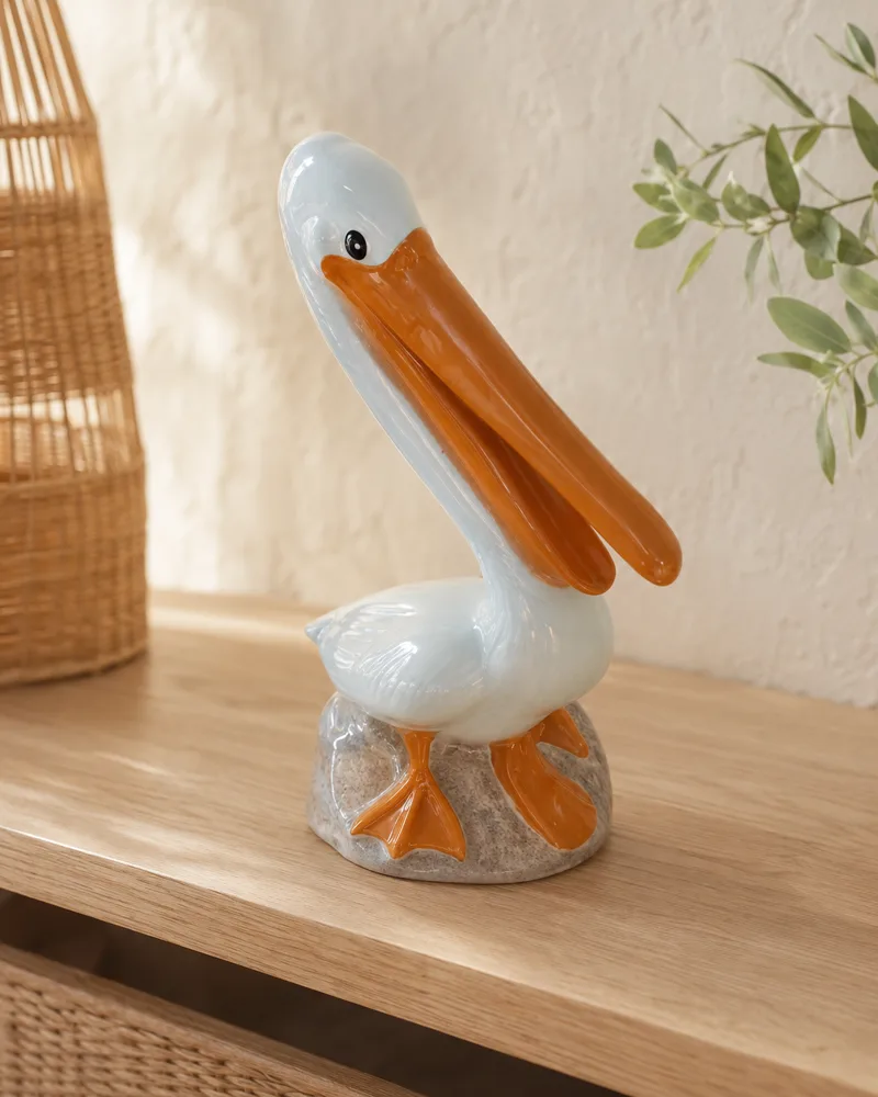 Glazed ceramic pelican figurine with an orange bill on a light oak console