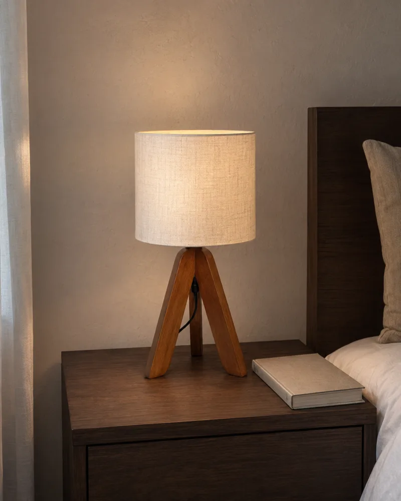 Small tripod table lamp with a linen shade glowing on a dark wood nightstand